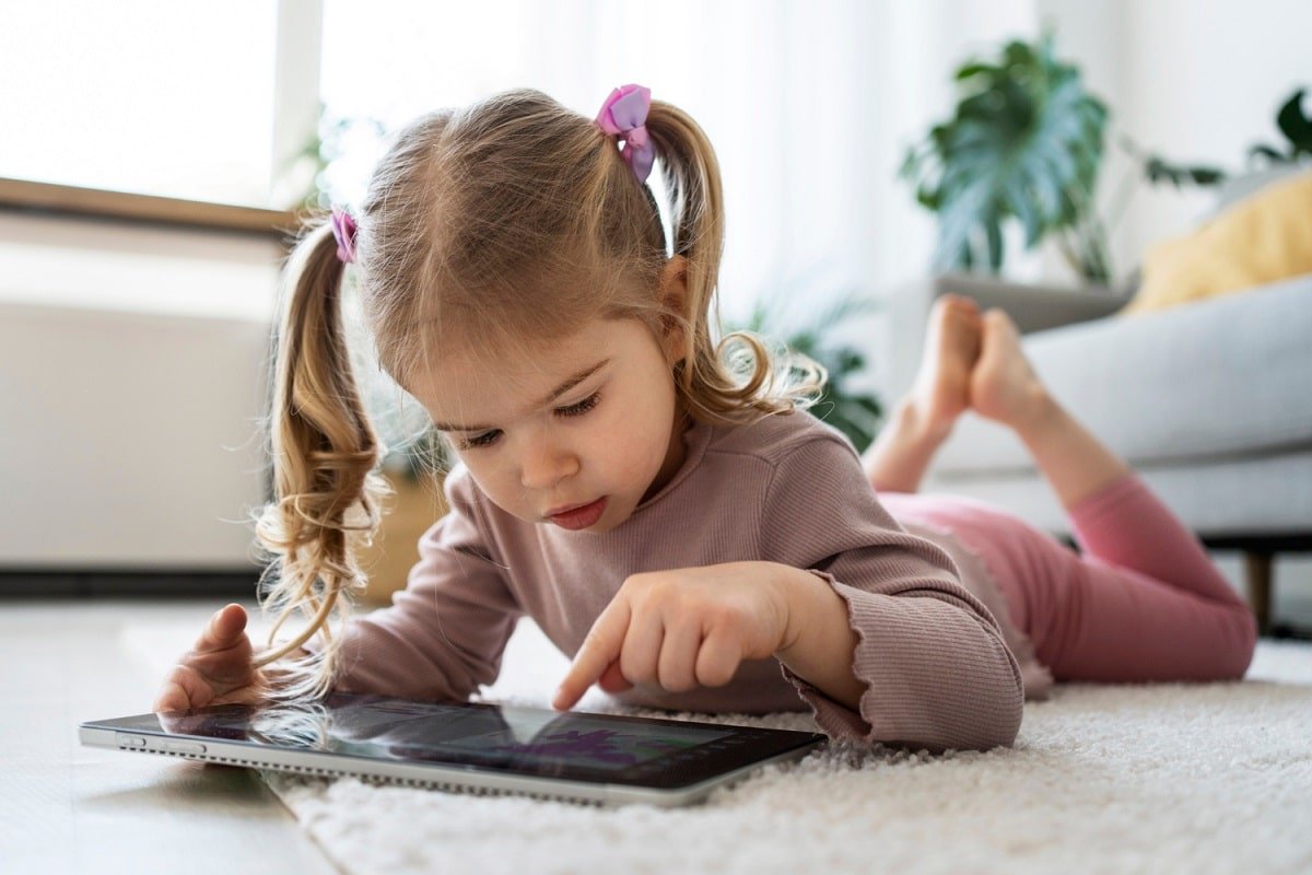 Une enfant avec des couettes utilise une tablette tactile allongée sur la moquette du salon