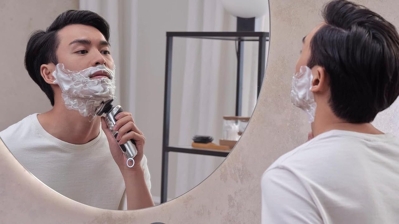 Un homme dans sa salle de bain qui se rase la barbe devant le miroir. Il utilise un rasoir de la marque Philips.