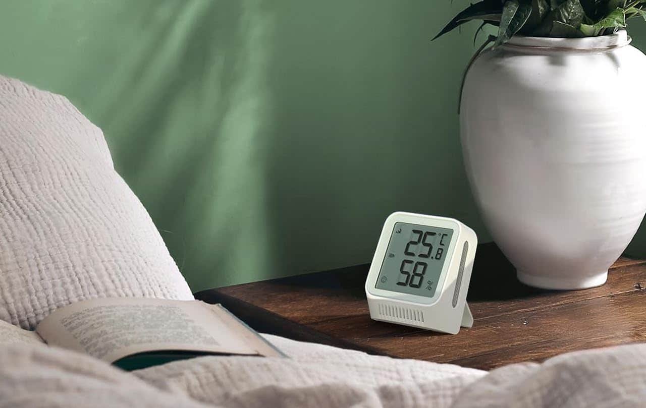 Un thermomètre connecté posé sur une table de chevet en bois avec un vase et des fleurs.